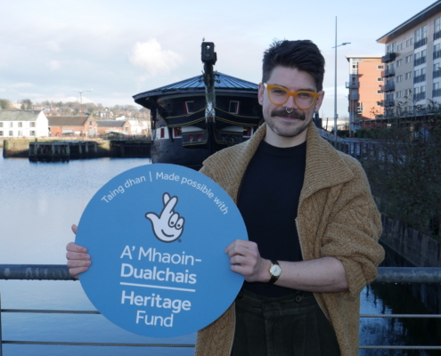 Marketing Agency Scotland - Cunningly Good Group - Image of Matthew Bellhuse Moran pictured in front of HMS Unicorn with a Heritage Fund lottery logo